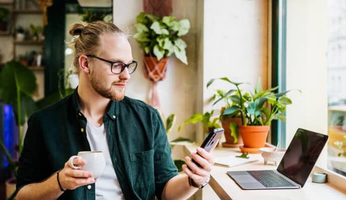 Muž se šálkem kávy používající mobilní telefon chráněný aplikací Norton 360 for Mobile.